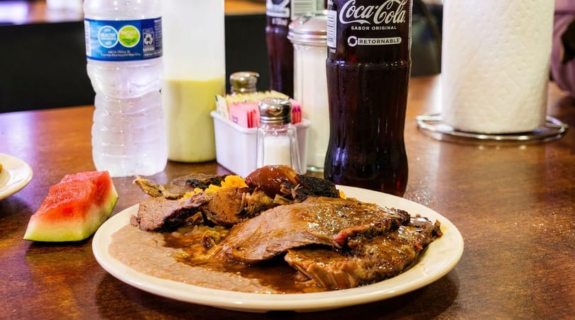 Braised beef with beans, watermelon, and drinks on a restaurant table with condiments.
