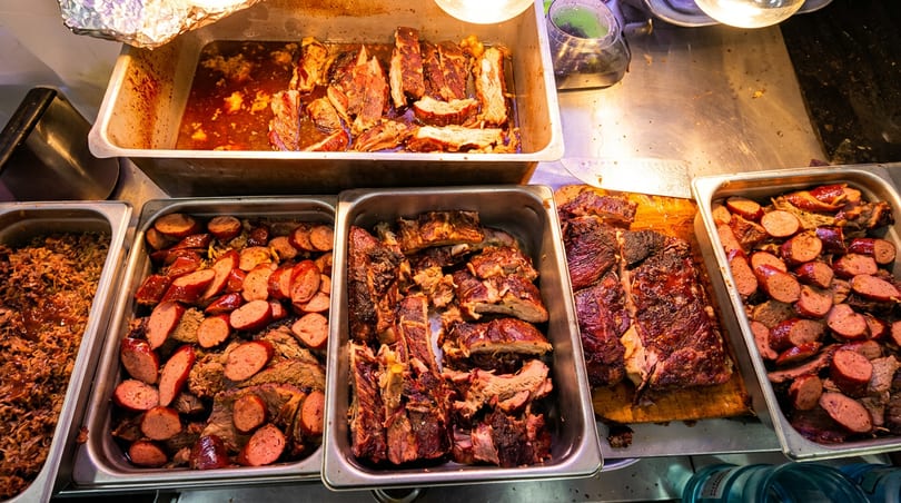 Hot buffet line with trays of BBQ ribs, pulled pork, and sliced sausage.