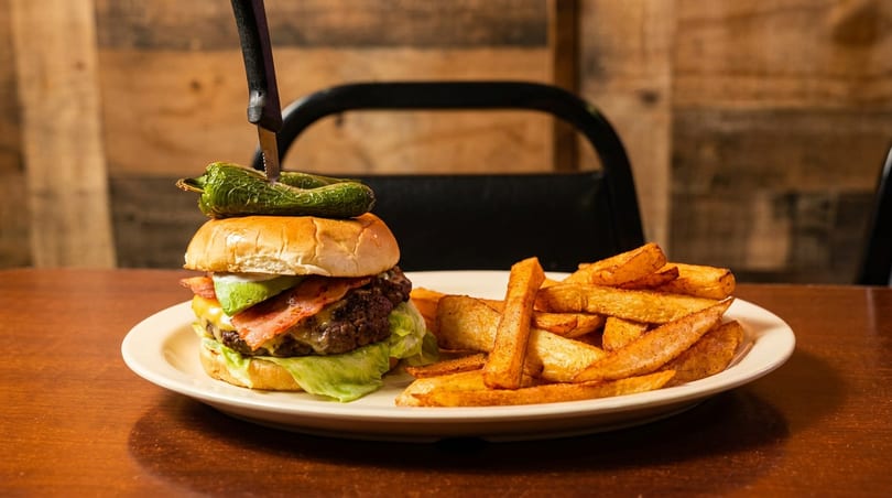 A large burger with bacon, avocado, and a pepper, skewered with a knife, alongside wedge fries.