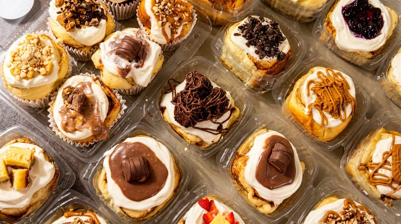 Assortment of gourmet cinnamon rolls with various toppings in individual plastic containers.